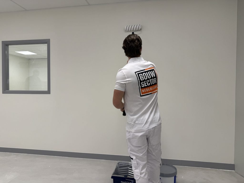 Schilder in wit werkpolo rolt een lichte wand in een lege ruimte, verfbak en emmer aan zijn voeten