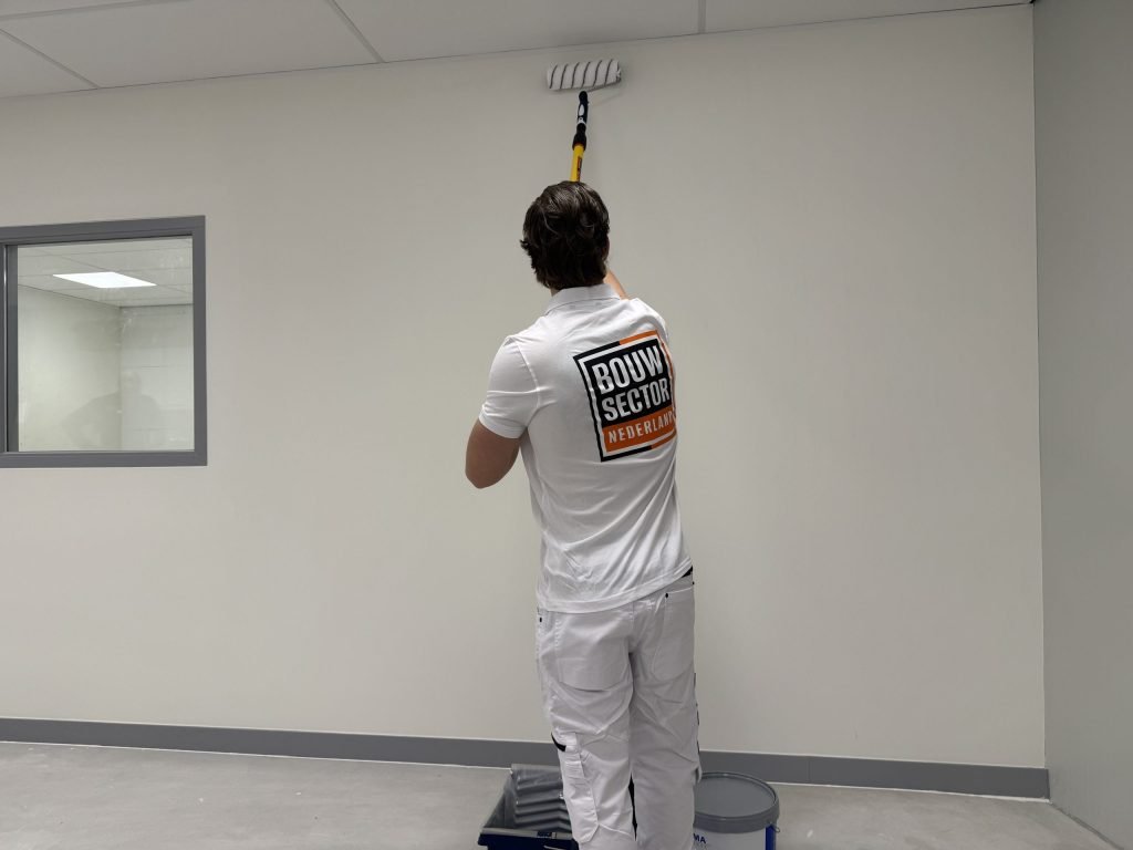 Schilder zet de roller hoog op een witte muur, met verfbak en emmer bij zijn voeten