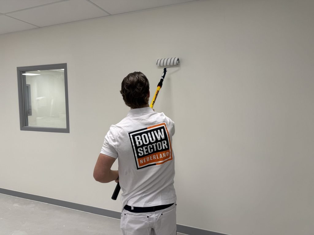 Schilder van Bouwsector Nederland rolt met een gele verlengsteel een witte wand naast een binnenraam