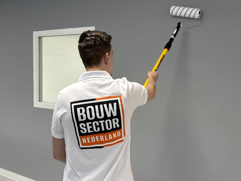Schilder met het Bouwsector-logo rolt een grijze muur, links een raam met witte omlijsting
