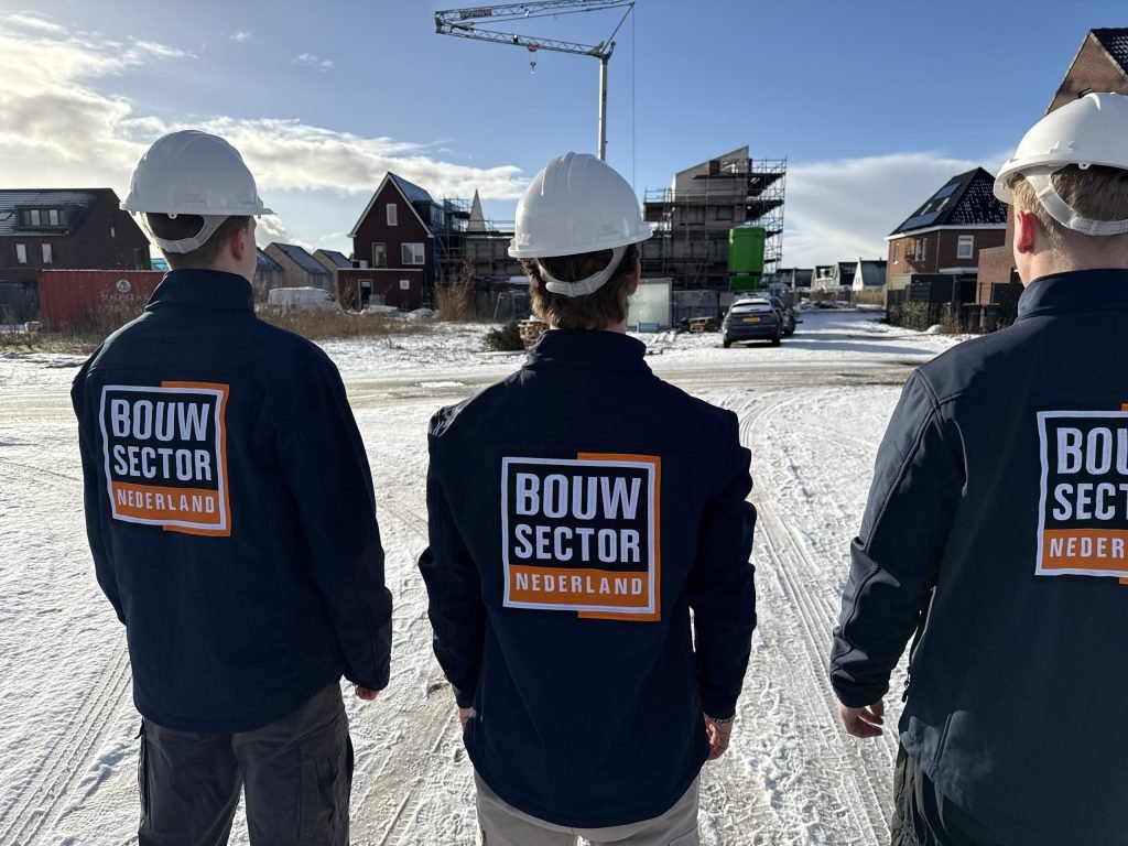 Drie vakmensen van Bouwsector Nederland met witte helm in een besneeuwde straat met nieuwbouw en een bouwkraan