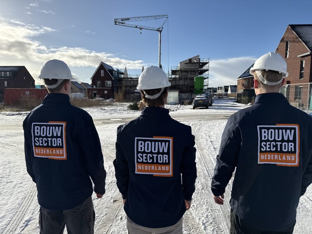 Drie vakmensen van Bouwsector Nederland met witte helm, van achteren gezien, in een besneeuwde nieuwbouwstraat met een bouwkraan